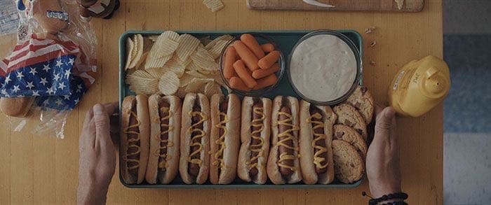 A still from the movie Weapons, showing a tray of 7 hot dogs, 4 chocolate chip cookies, a pile of potato chips, a bowl of carrots, and some sort of white dip (ranch?), which two gay men are about to enjoy as dinner