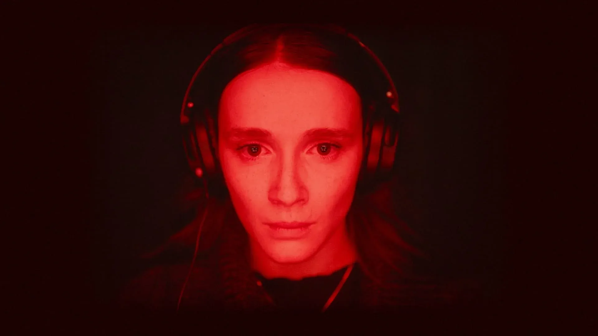 A still from Red Rooms, showing the main character, Kelly-Anne, her face lit red by the glow of her computer screen, wearing headphones and watching horrific gore, on her face an inscrutable but almost peaceful expression